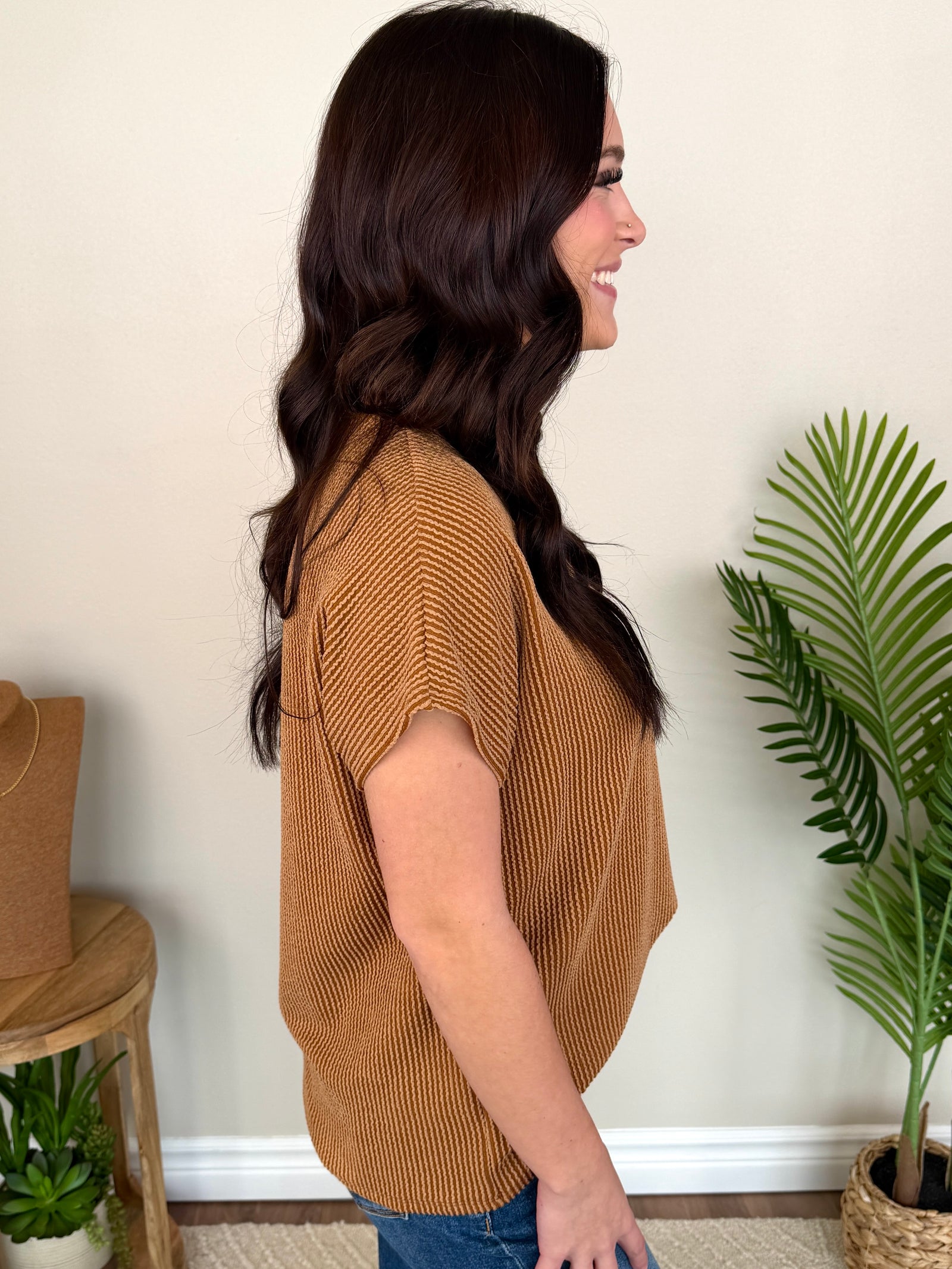 A woman with long dark hair smiling, wearing a short-sleeve ribbed knit top in a warm camel brown color, paired with blue jeans, standing in front of a beige wall with a green potted plant on the right.