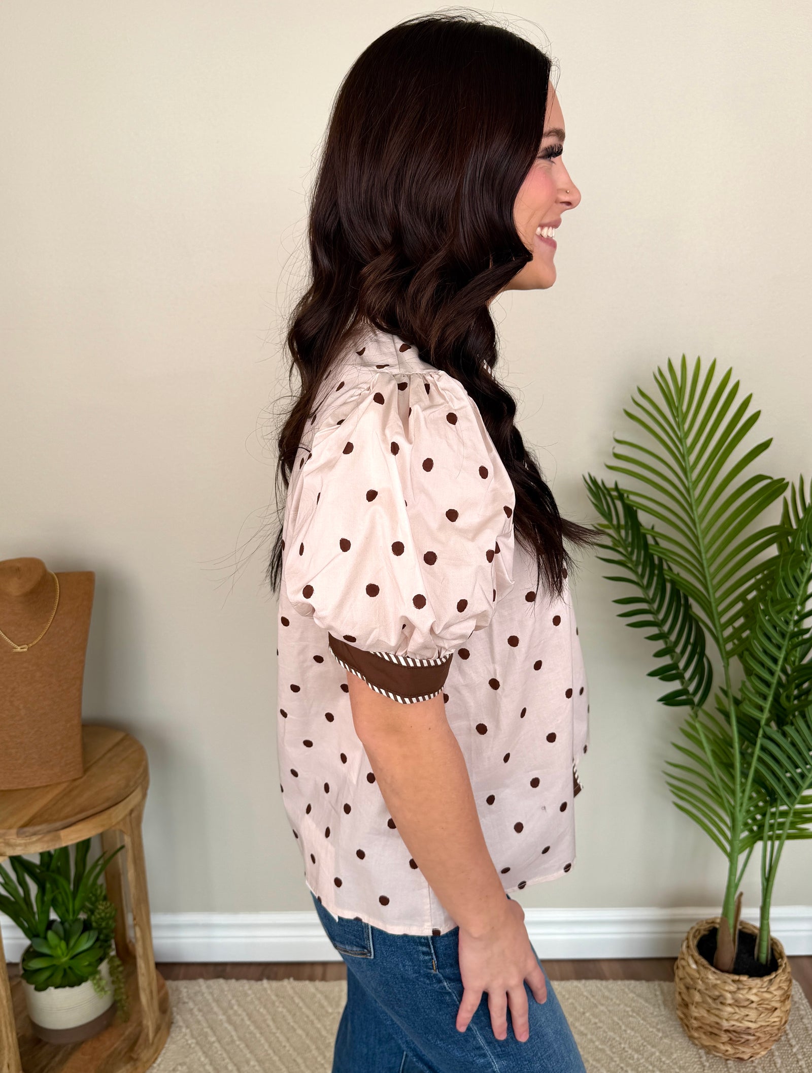 A smiling woman with long dark hair stands indoors wearing a light blush pink short-sleeve blouse with brown polka dots and brown trim, paired with blue jeans. The background shows a beige wall with a potted palm and a small wooden side table.