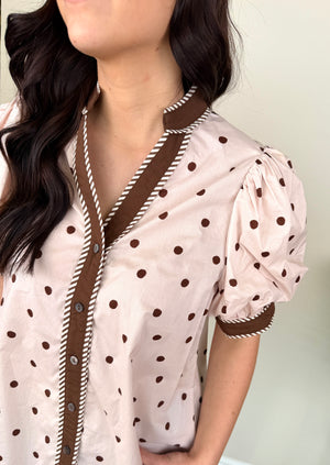 A close-up of a woman wearing a light pink short-sleeve blouse with brown polka dots and contrasting brown trim with white rope-style edging along the collar, placket, and sleeve cuffs.