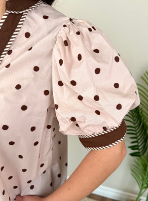 A close-up of a person wearing a light pink puff-sleeve top with dark brown polka dots and striped trim, shown against an indoor background with a plant.