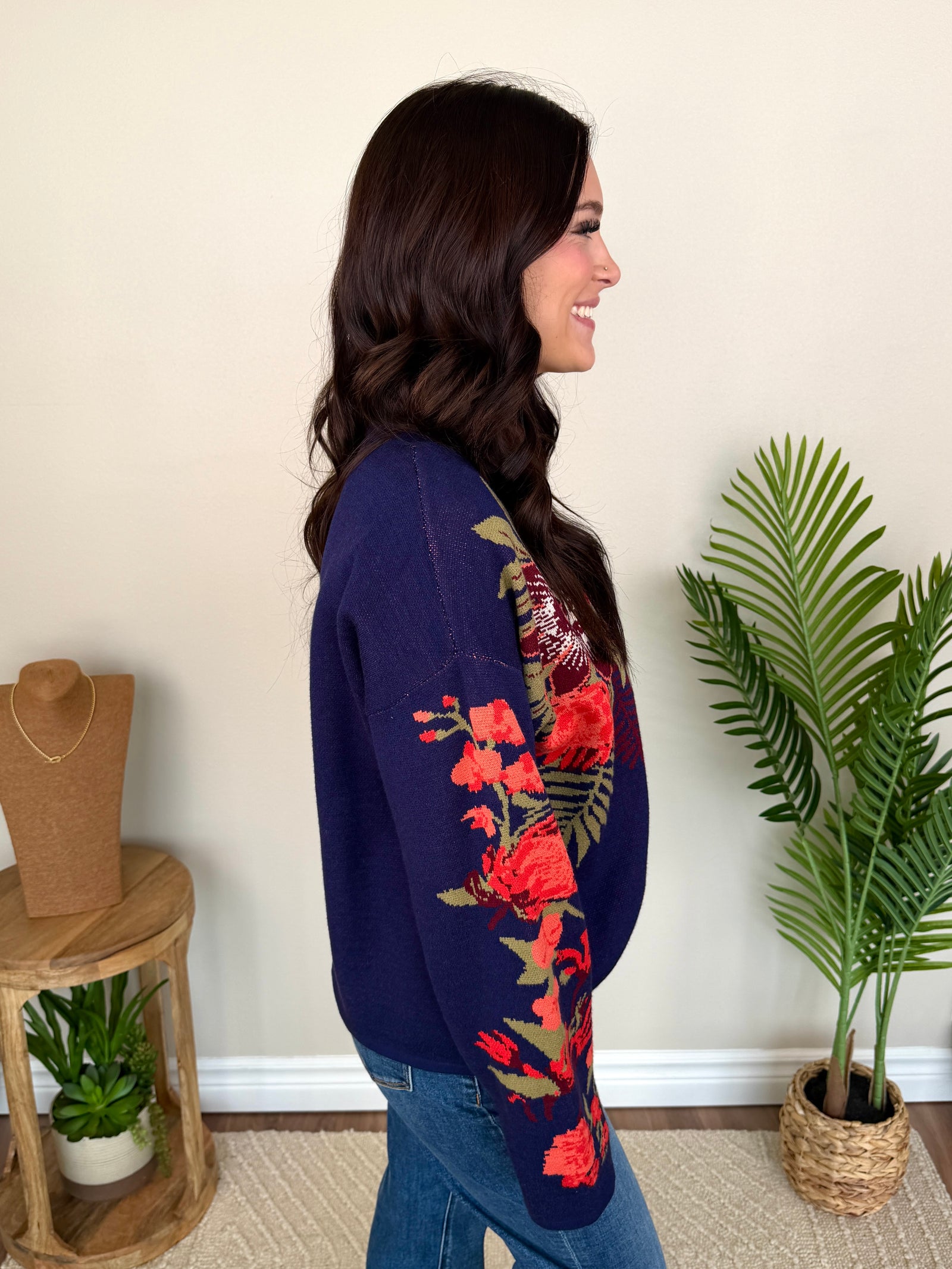 A woman smiling while wearing a navy blue mock-neck sweater with red and orange floral patterns and green leaves, paired with blue jeans, standing in an indoor setting with a plant and wooden furniture.
