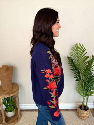 A woman wearing a navy blue knit sweater with red and green floral embroidery on the sleeves, standing in a room with indoor plants and a jewelry display stand.
