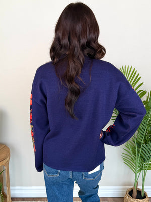 A woman viewed from behind wearing a navy blue knit sweater with colorful red, orange, and yellow graphic details on the sleeves, paired with blue jeans, standing indoors next to a potted plant.