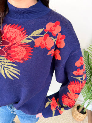 A close-up of a person wearing a navy blue knit sweater with large red and orange floral patterns and green leaves across the chest and sleeves, paired with blue jeans in an indoor setting with a plant visible.