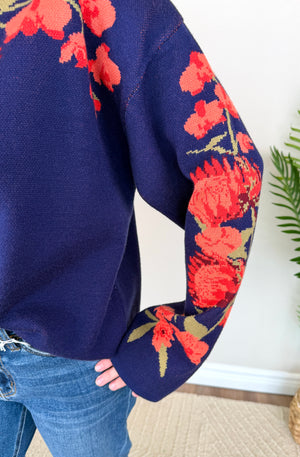 A close-up of a person wearing a navy blue knit sweater with a vibrant red and orange floral pattern on the sleeves and upper chest, paired with blue jeans.