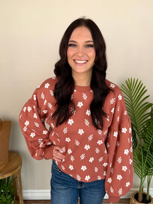 A smiling woman with long dark hair wearing a terracotta-colored long-sleeve sweatshirt with a white floral and leaf pattern, paired with blue jeans, standing in an indoor setting with a beige wall and green plants.
