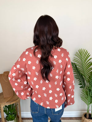 A woman viewed from behind wearing a dusty rose long-sleeve blouse with a white floral and leaf print, paired with blue jeans in an indoor setting with a potted plant and wooden stool.