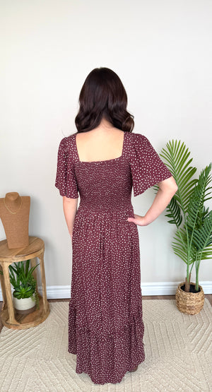A woman stands with her back to the camera wearing a dark burgundy maxi dress with a small white floral pattern. The dress features short puff sleeves, a square neckline, and a tiered ruffled hem.