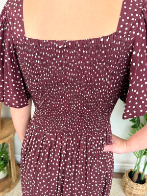 A close-up of a person wearing a smocked dress in burgundy with white polka dots. The dress features a square neckline, short puff sleeves, and a gathered skirt, shown in a home setting with plants in the background.