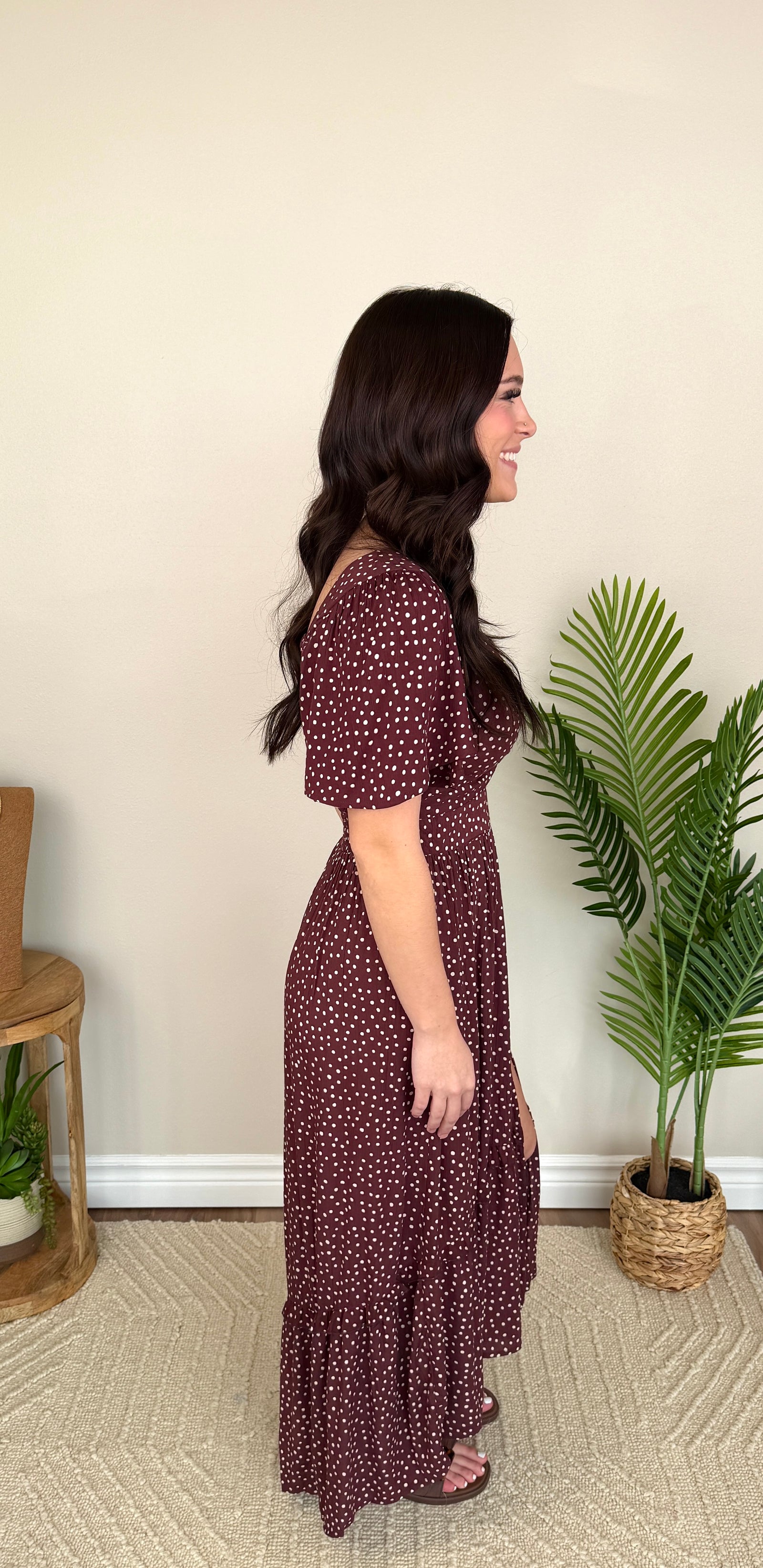 A woman modeling a short-sleeve wrap maxi dress with a small white floral print on a dark burgundy background, featuring a keyhole cutout at the waist and a thigh-high slit, standing in a styled indoor setting with a potted plant and a wooden side table.