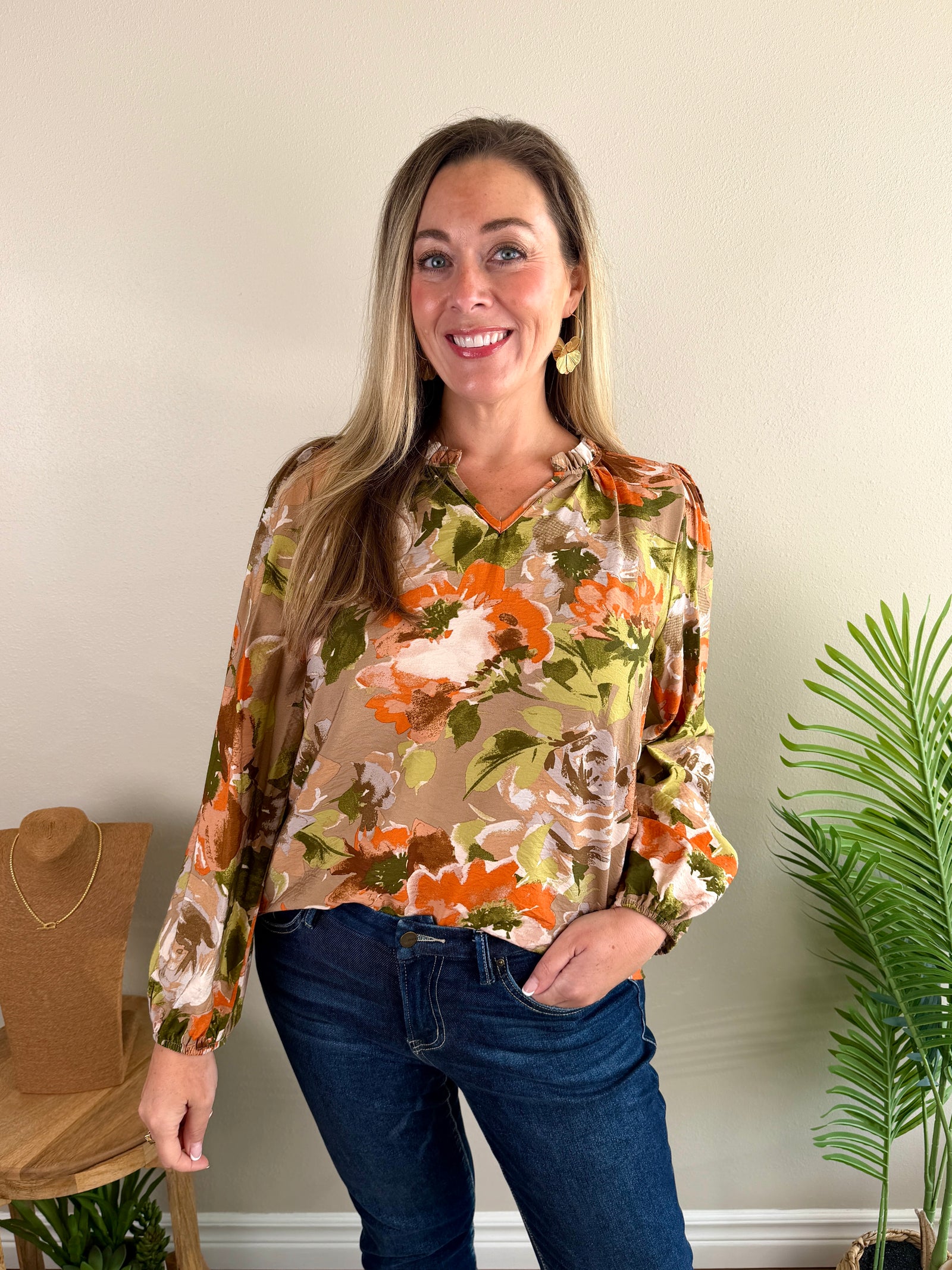 A woman wearing a beige long-sleeve blouse with a vibrant orange, green, and white floral print, paired with dark blue jeans, standing in a room with a beige wall, a potted plant, and a wooden stand.