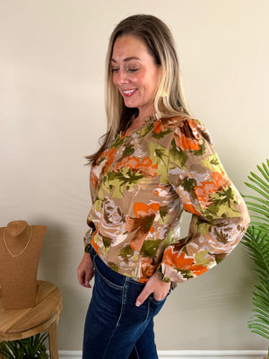 A woman modeling a long-sleeve blouse with a retro floral pattern in orange, olive green, and beige, paired with dark blue jeans, standing in a room with a beige wall.