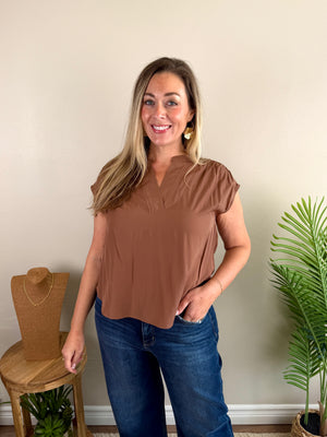 A woman modeling a short-sleeve brown top with a split neckline, paired with blue jeans, standing in a room with a beige wall, a wooden stool, and a potted plant.