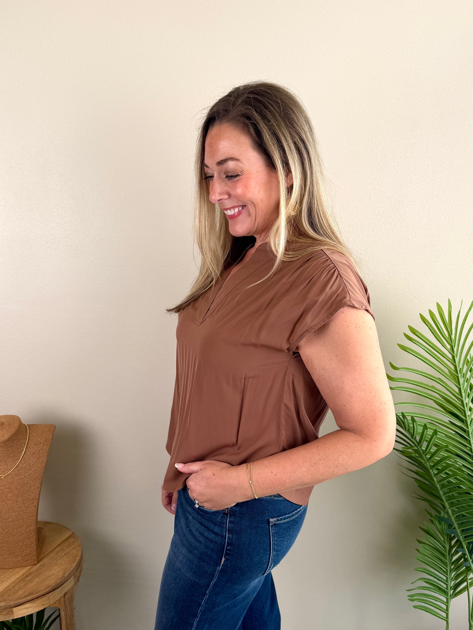 A woman modeling a short-sleeve brown top with a split neckline, paired with blue jeans, standing in a room with a beige wall, a wooden stool, and a potted plant.