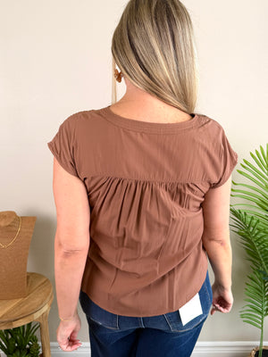 A woman viewed from the back wearing a short-sleeve brown top with a gathered, smocked back panel and blue jeans, standing indoors against a light beige wall with a plant and wooden stool visible.