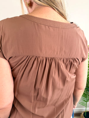 A close-up back view of a woman wearing a short-sleeve dusty brown top with a gathered waist seam, set against a light indoor background.