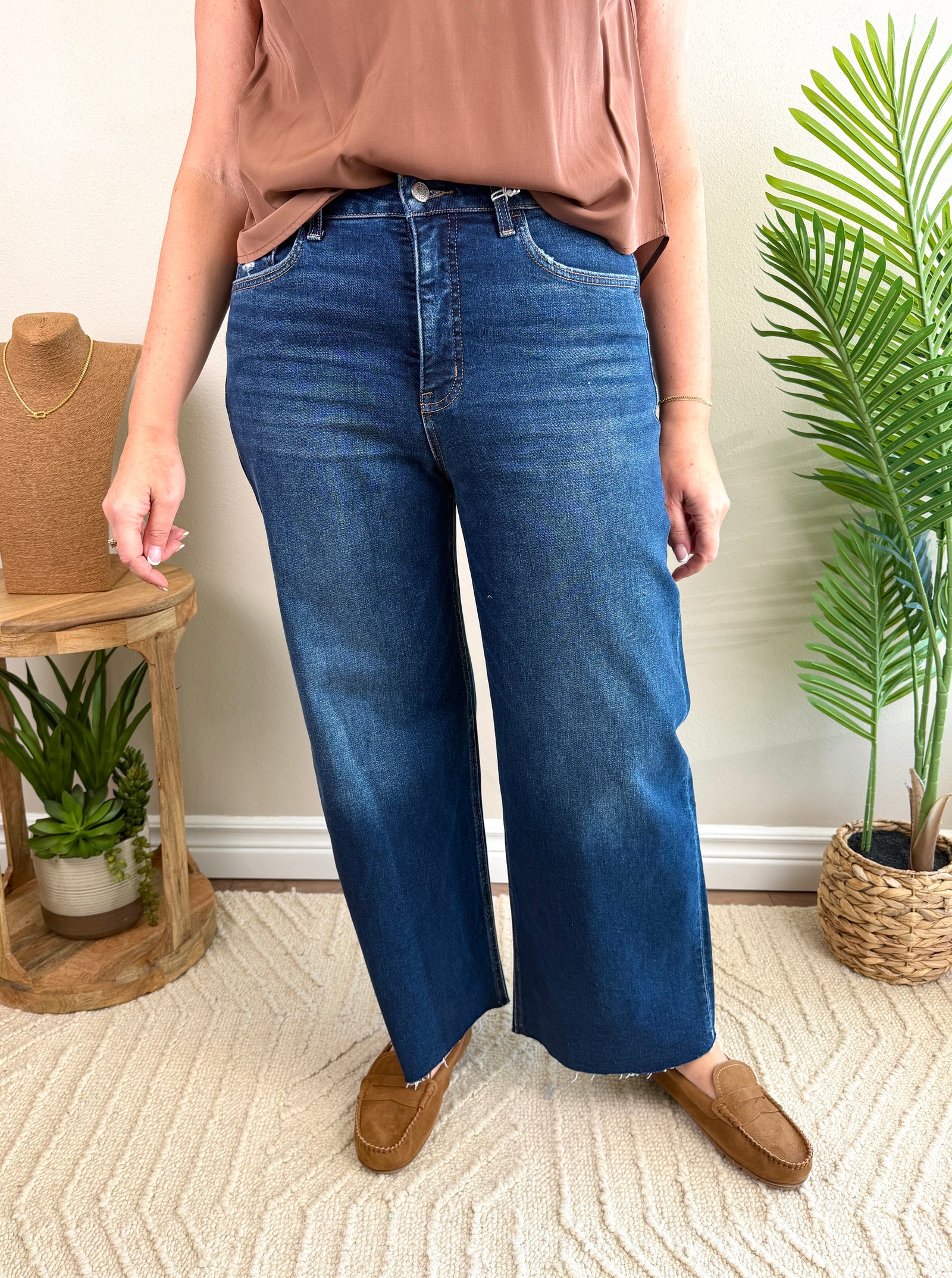 A person wearing high-waisted, wide-leg blue jeans with a tan top and brown slip-on shoes, standing in a styled indoor room with plants and a textured rug.