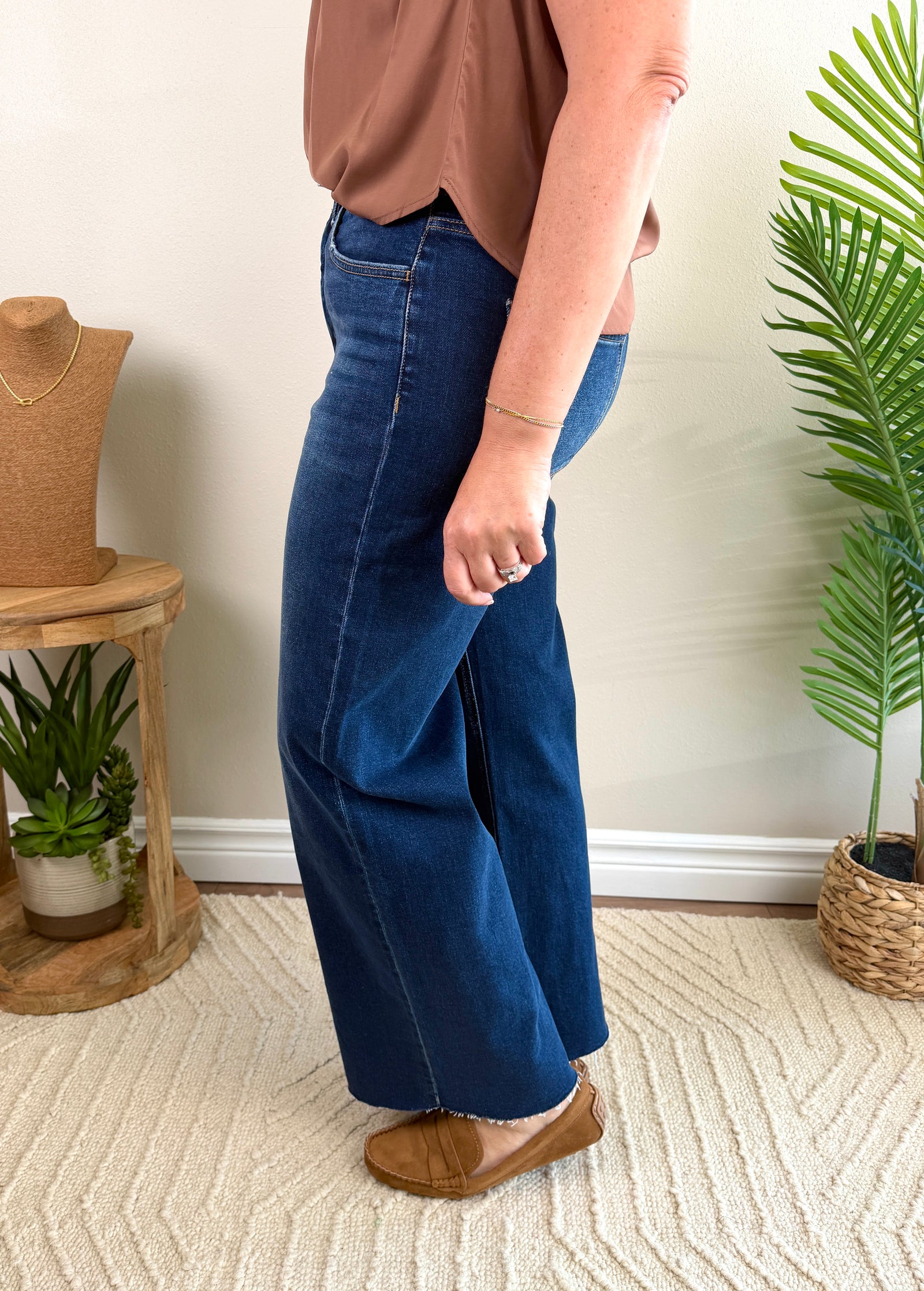 A person wearing high-waisted, wide-leg blue jeans with a tan top and brown slip-on shoes, standing in a styled indoor room with plants and a textured rug.