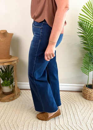 A person wearing dark blue wide-leg denim jeans, a brown sleeveless top, and tan mule flats, standing in a styled indoor room with a beige wall, patterned rug, and potted plants.