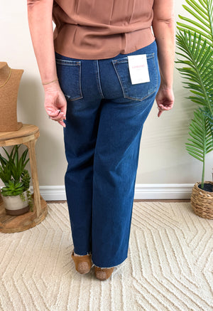 A person shown from the back wearing blue jeans with a white paper tag attached to the back pocket, standing indoors on a textured cream rug with plants and furniture in the background.