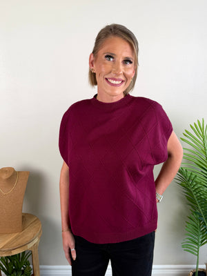 A woman wearing a short-sleeve crewneck sweater in deep burgundy with a diamond knit pattern, standing in front of a white wall with a plant and jewelry stand visible.
