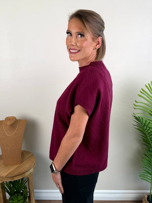 A woman wearing a short-sleeve mock neck sweater in deep burgundy with a subtle diamond knit pattern, standing in a room with a light beige wall, a wooden jewelry stand, and a green plant.
