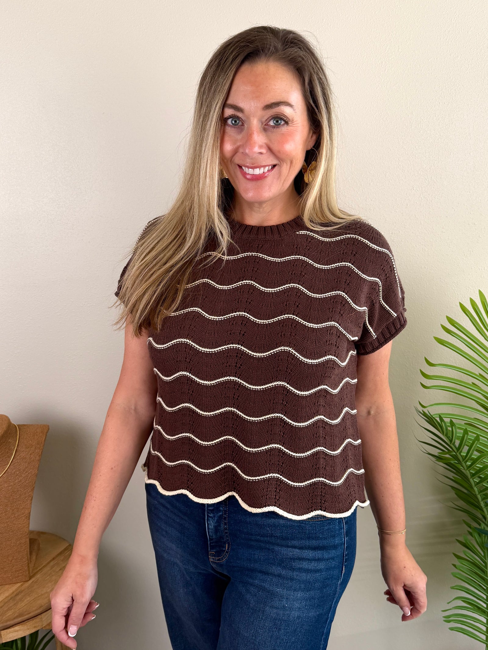 A woman wearing a brown short-sleeve knit top with wavy cream stripes and blue jeans, standing against a beige wall with a plant and hat visible in the background.