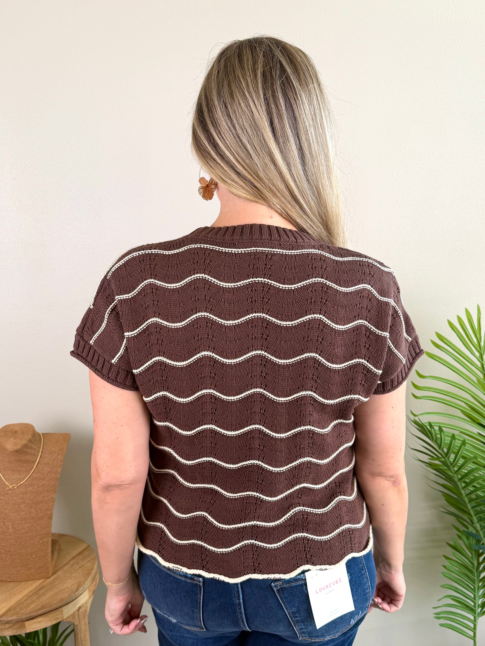 A woman wearing a brown short-sleeve knit top with wavy cream stripes and blue jeans, standing against a beige wall with a plant and hat visible in the background.