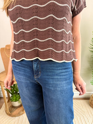 A person wearing a brown short-sleeve knit top with cream wavy horizontal stripes and blue jeans, standing indoors near a chair and plant.