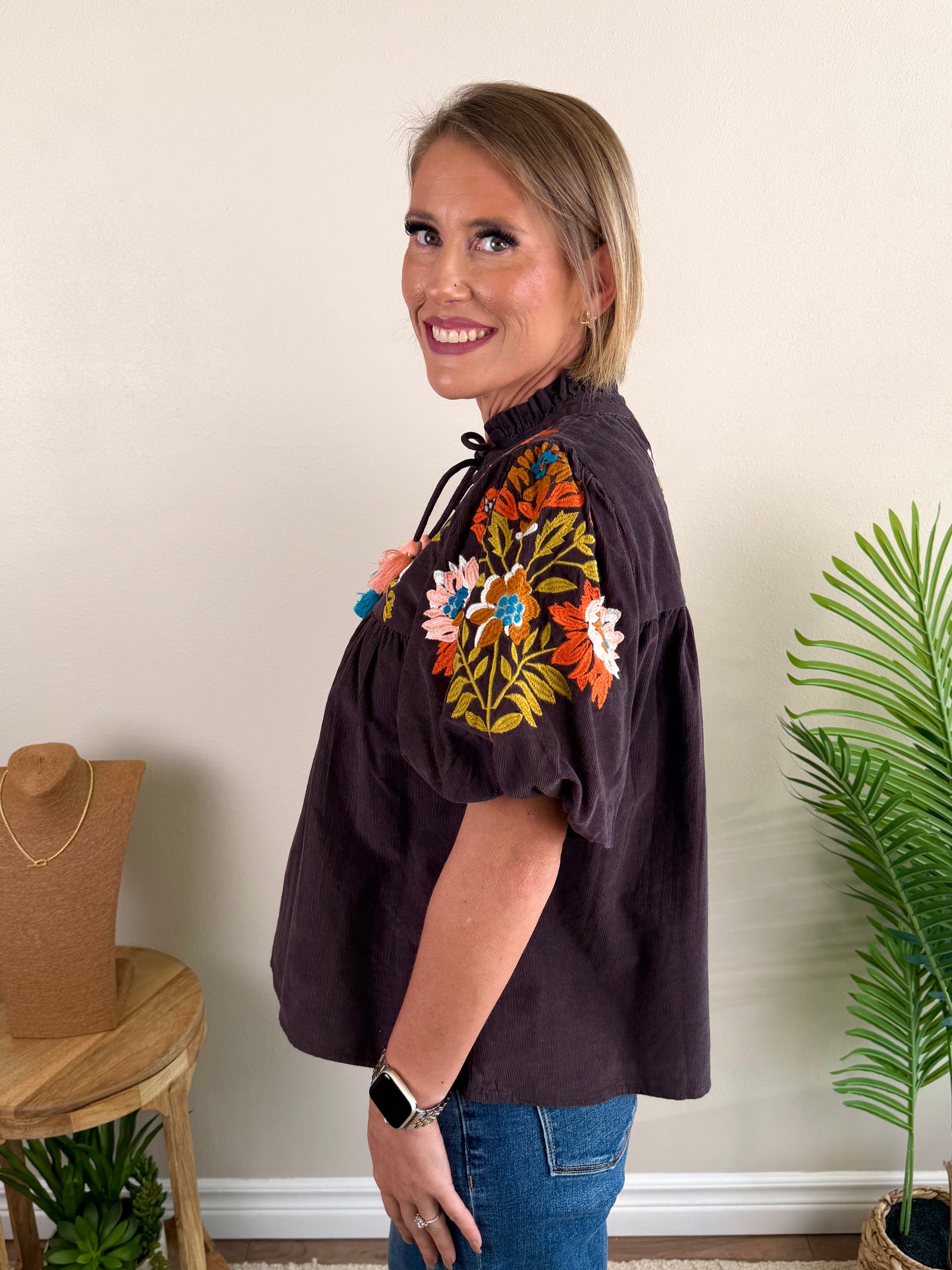 A woman wearing a dark brown embroidered blouse with colorful floral patterns and blue tassel ties, paired with blue jeans, standing in front of a beige wall.