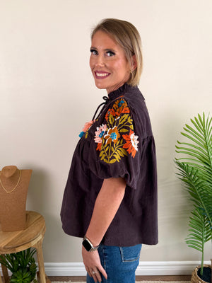 A woman modeling a dark brown short-sleeve blouse with colorful floral embroidery on the shoulder, paired with blue jeans, standing in a room with a beige wall, a plant, and a jewelry stand.
