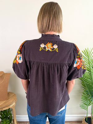 A woman viewed from behind wearing a dark brown short-sleeve blouse with colorful floral embroidery on the back neckline and sleeves, paired with blue jeans in an indoor setting.