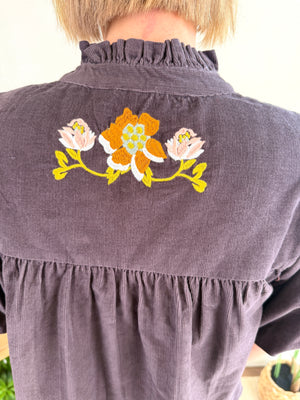A close-up back view of a person wearing a dark brown corduroy blouse with a ruffled high neck and embroidered flowers on the upper back.