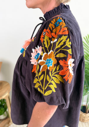 A close-up side view of a woman wearing a dark brown corduroy blouse with vibrant multicolor floral embroidery on the sleeve and chest. The blouse features a ruffled high neckline and drawstrings.