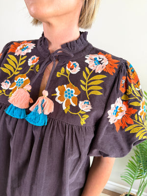 A close-up of a woman wearing a dark brown blouse with colorful floral embroidery, peach tassels, and a keyhole tie neckline.