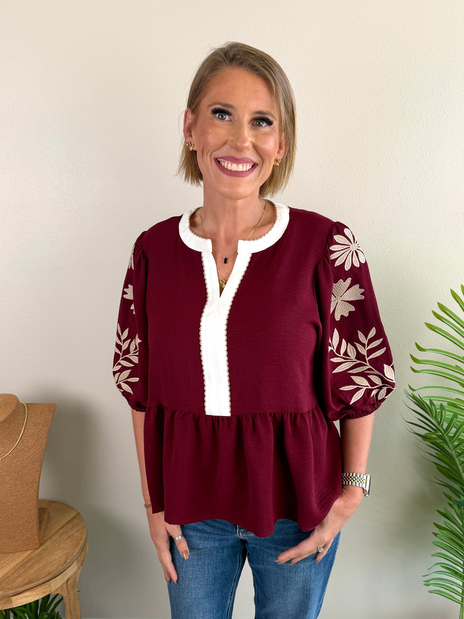 A woman wearing a burgundy peplum blouse with white floral embroidery on the sleeves and a white V-neck trim, paired with blue jeans, standing against a plain beige wall.