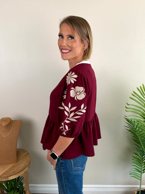 A woman wearing a burgundy peplum blouse with cream floral embroidery on the sleeves, paired with blue jeans, standing in a room with a beige wall, a wooden stool, and a green plant.