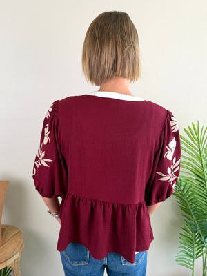 A woman viewed from behind wearing a burgundy blouse with white floral embroidery on the sleeves and a white collar, paired with blue jeans against a plain indoor wall.