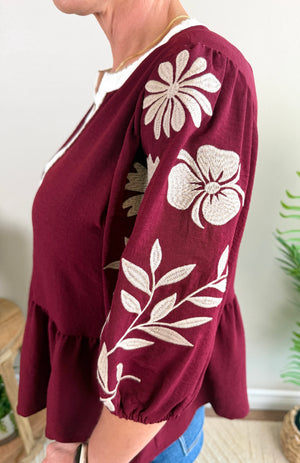 A woman wearing a burgundy peplum blouse with cream floral embroidery on the sleeves, paired with blue jeans.