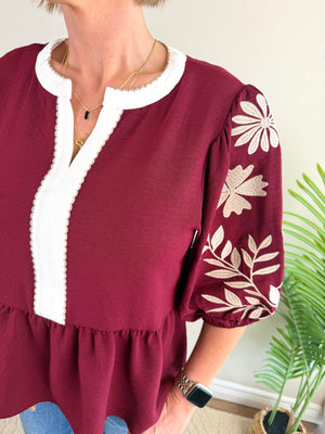 A woman wearing a burgundy peplum blouse with white embroidered floral details on the sleeves and a white scalloped trim around the neckline and front placket.