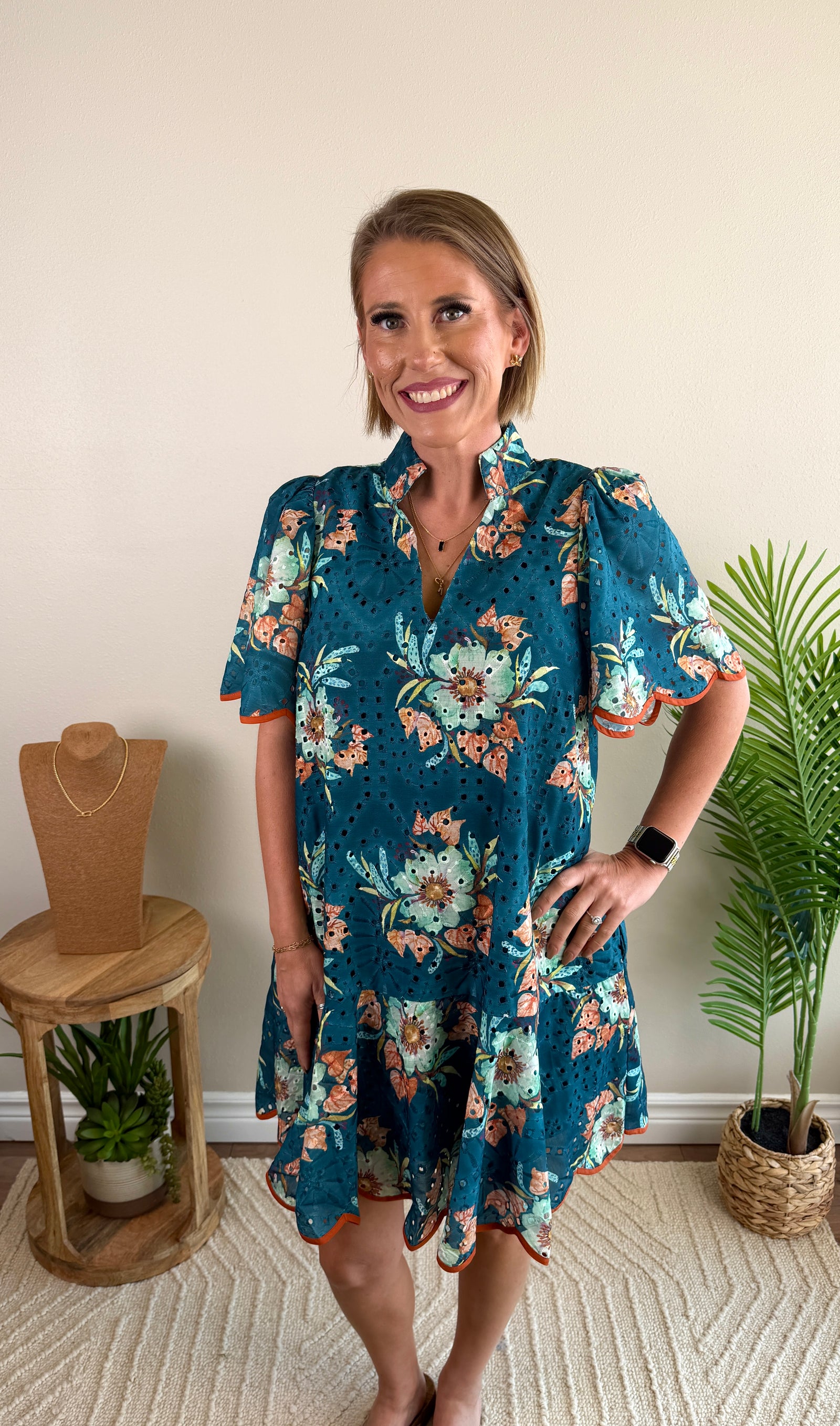 A woman modeling a short-sleeve teal floral dress with orange and white accents, standing in a brightly lit indoor room with a beige wall, a small wooden stand, and a potted plant.