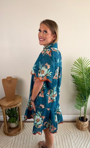 A woman modeling a teal blue floral short-sleeve tunic dress with orange ruffle trim, standing in a room with a beige wall, a potted plant, and a wooden stand.