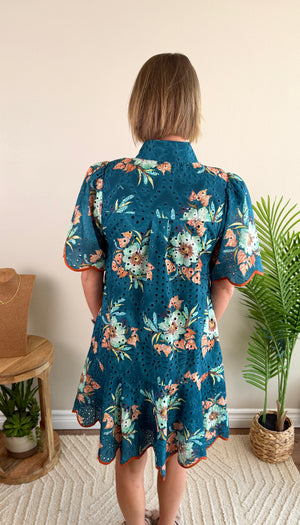 A woman viewed from behind wearing a teal blue short-sleeve eyelet dress with a colorful floral print, standing in a room with a beige wall, potted plants, and a small wooden side table.