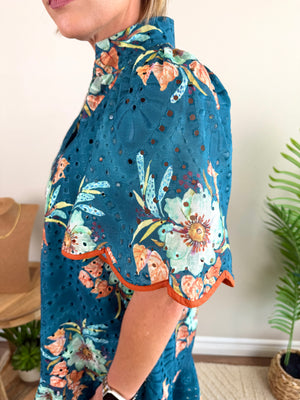 A close-up side view of a woman wearing a teal eyelet blouse with a colorful floral and butterfly print, featuring orange scalloped trim on the sleeve.