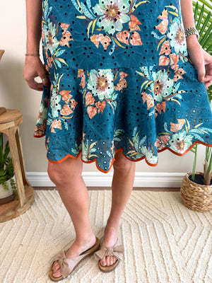 A person wearing a teal blue floral eyelet dress with orange and peach flowers, paired with beige slide sandals, standing on a textured cream rug in a home setting.